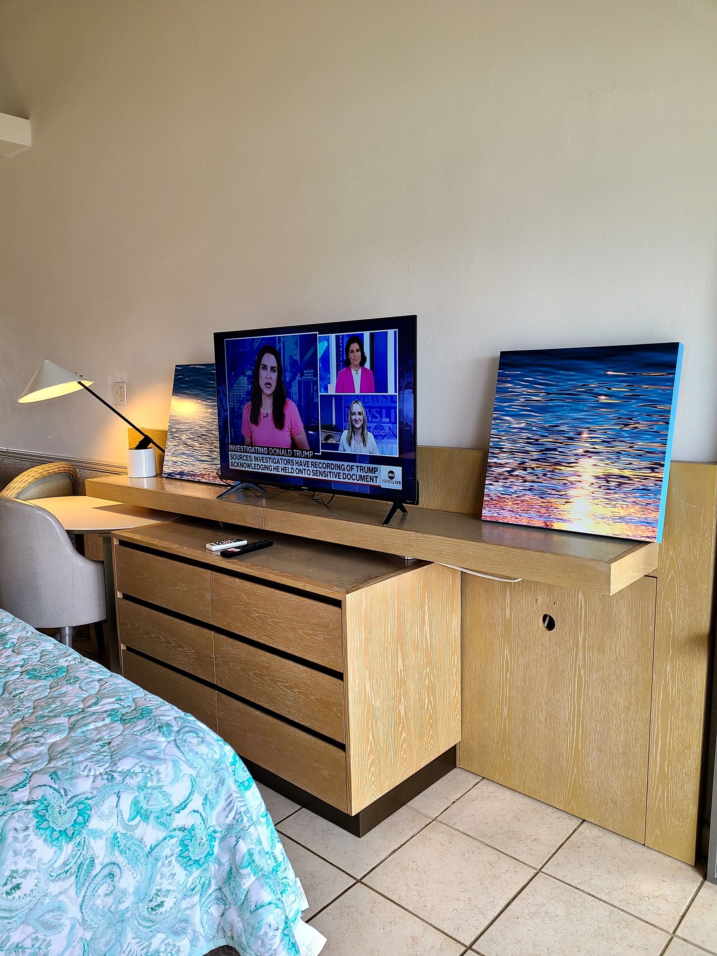 A hotel room with a wooden desk, a television displaying a news broadcast, a painting of a seascape, and a bed with a patterned bedspread.