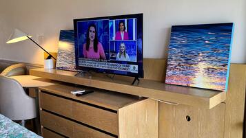 A hotel room with a wooden desk, a television displaying a news broadcast, a painting of a seascape, and a bed with a patterned bedspread.