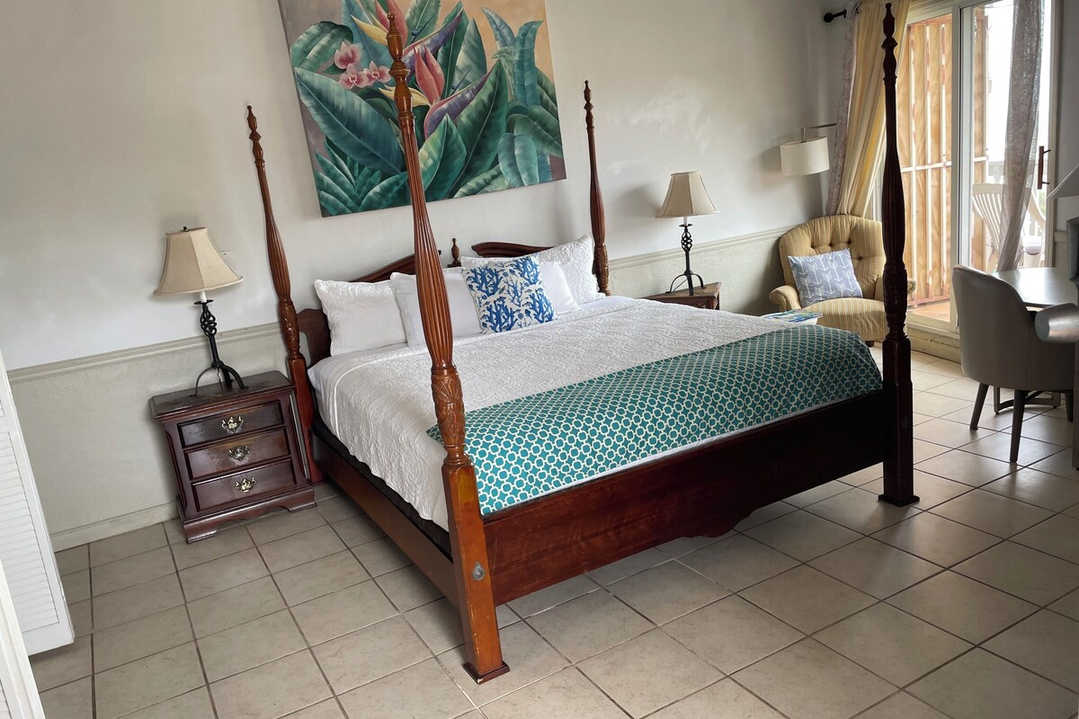 A four-poster bed with a patterned bedspread, two bedside tables with lamps, a chair, and a large botanical painting on the wall.