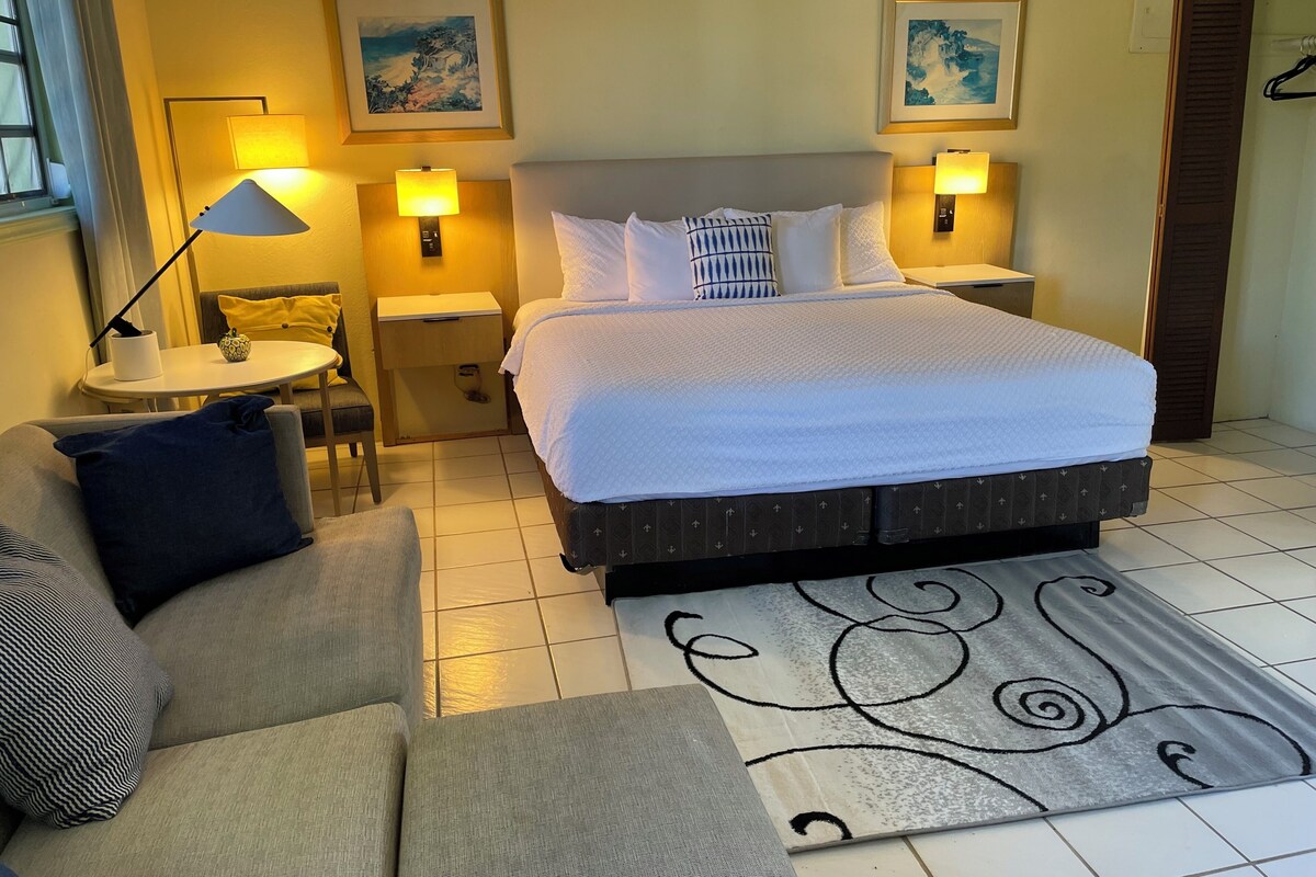 A hotel room with a bed, two bedside lamps, a small table, a chair, and a sofa.