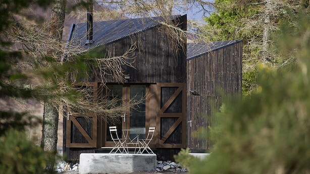 A rustic cabin with a wooden exterior, a large window, and a patio with chairs. - Laggan Bothies (Spean Bridge)