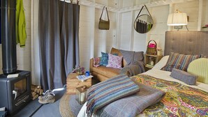 A cozy room with a wooden interior, a bed with colorful bedding, a small table with a lamp, and a fireplace. - Laggan Bothies (Spean Bridge)