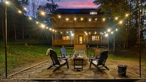 Una cabaña de madera con un patio de piedra, mobiliario exterior e iluminación con luces colgantes.