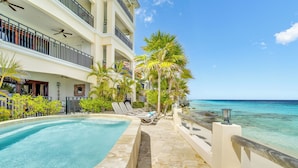A resort pool area with a clear view of the ocean, a multi-story building with balconies, and palm trees.