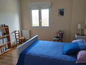 Un dormitorio con cama, una silla, una estantería, una ventana y una mesita de noche.