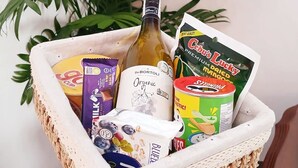 A woven basket containing various food items and a bottle of wine, placed on a wooden surface with a potted plant in the background.