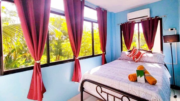 A bedroom with a bed, curtains, an air conditioner, and a view of greenery.
