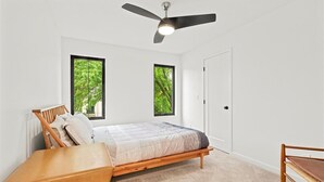 A bedroom with a bed, a wooden bench, a ceiling fan, and two windows with a view of trees. - Modern 4 Bed/3.5 Bath Farmhouse Steps to Downtown CF! (Cedar Falls)
