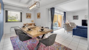 Ein modernes Wohnzimmer mit Essbereich, mit einem Glastisch, Korbstühlen, einem blauen Sofa und einem Fernseher.