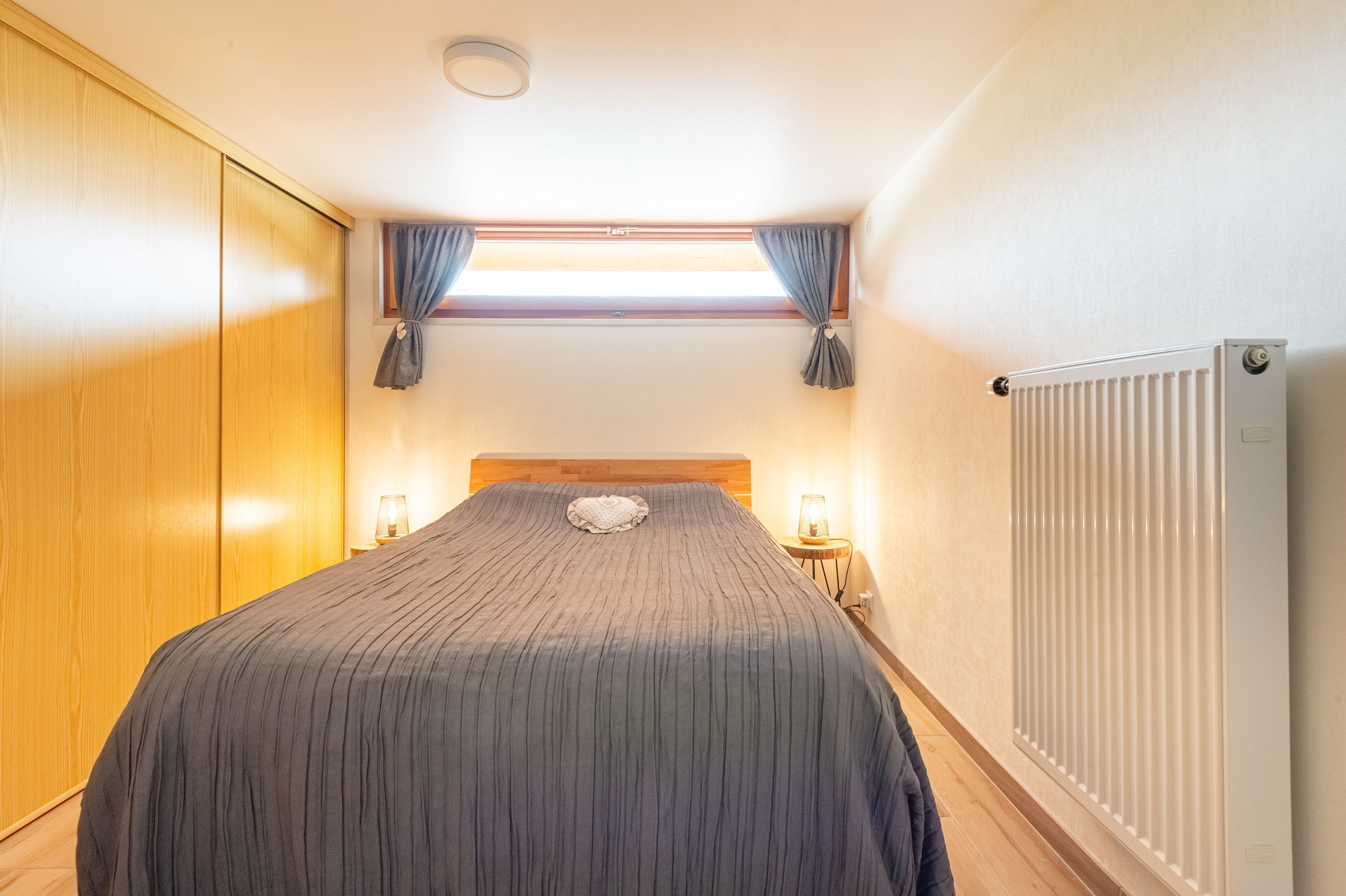 A bedroom with a bed, a window with curtains, a radiator, and a nightstand with a lamp.