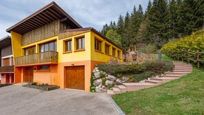 Un edificio amarillo de dos pisos con un balcón de madera, una escalera de piedra y un cerro cubierto de bosque al fondo.