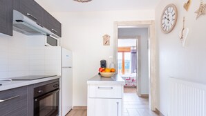 Una cocina moderna con armarios oscuros, una encimera blanca y un horno empotrado. Hay una pequeña isla con un cuenco de fruta y una cafetera. Un reloj y elementos decorativos están en la pared.