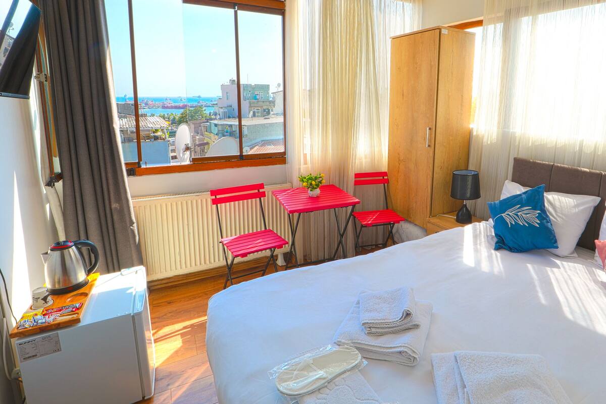 A hotel room with a bed, a table with chairs, a radiator, a kettle, and a view of the city.