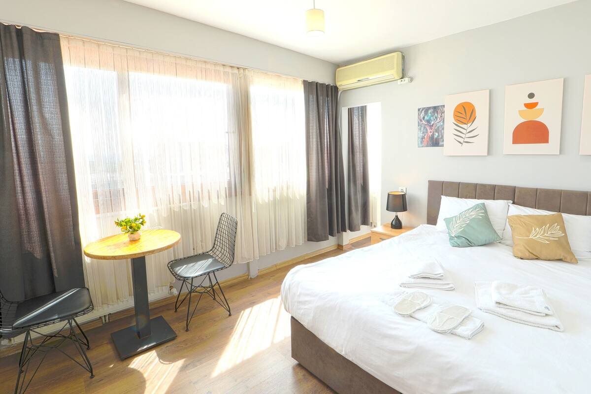 A hotel room with a bed, a round table with a plant, a chair, and artwork on the wall.