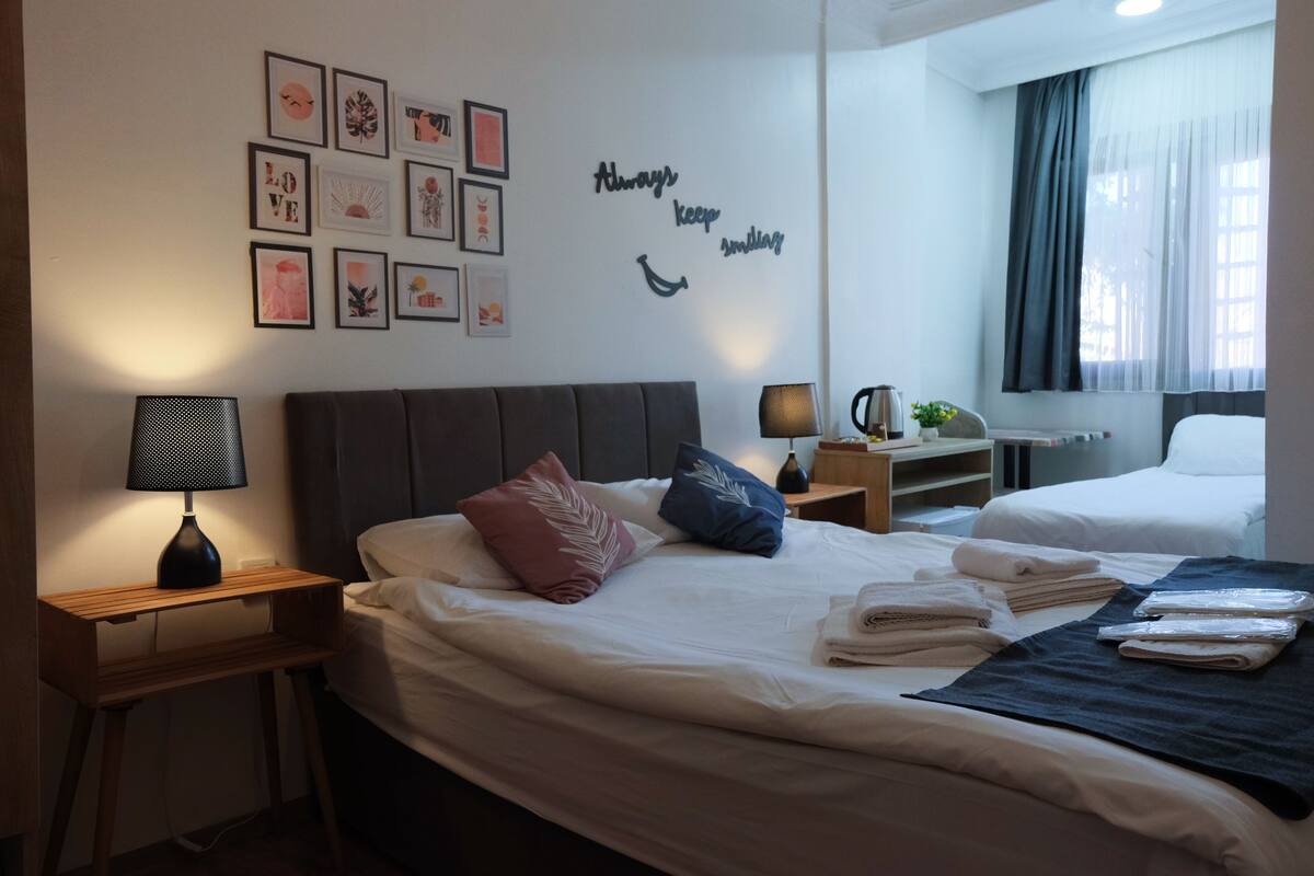 A hotel room with two beds, a bedside table with a lamp, a wall with framed artwork, and a window with curtains.