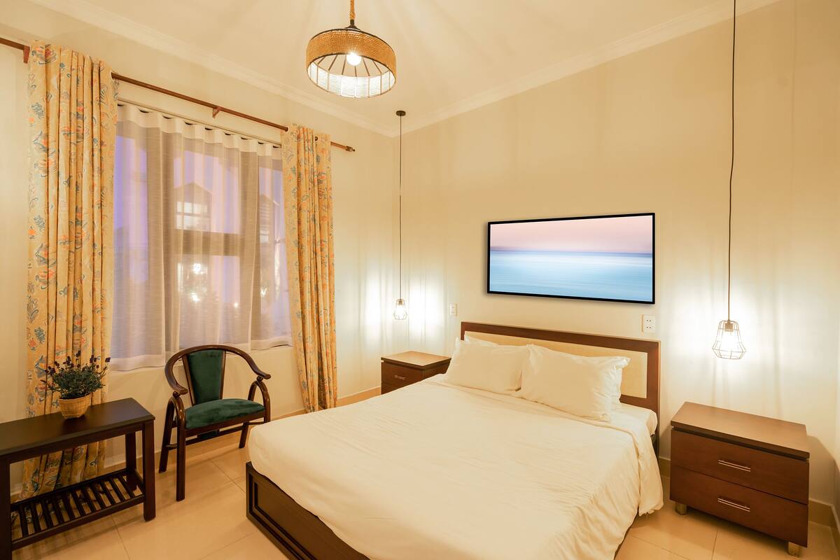 A neatly arranged bedroom with a bed, bedside tables, a chair, a TV, and a window with curtains.