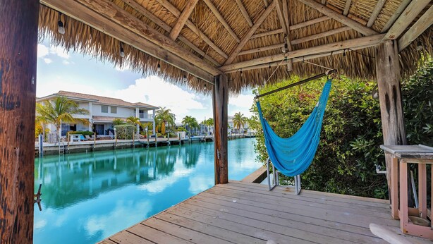 Terraza de madera con hamaca, piscina y vista a un resort con edificios y palmeras.