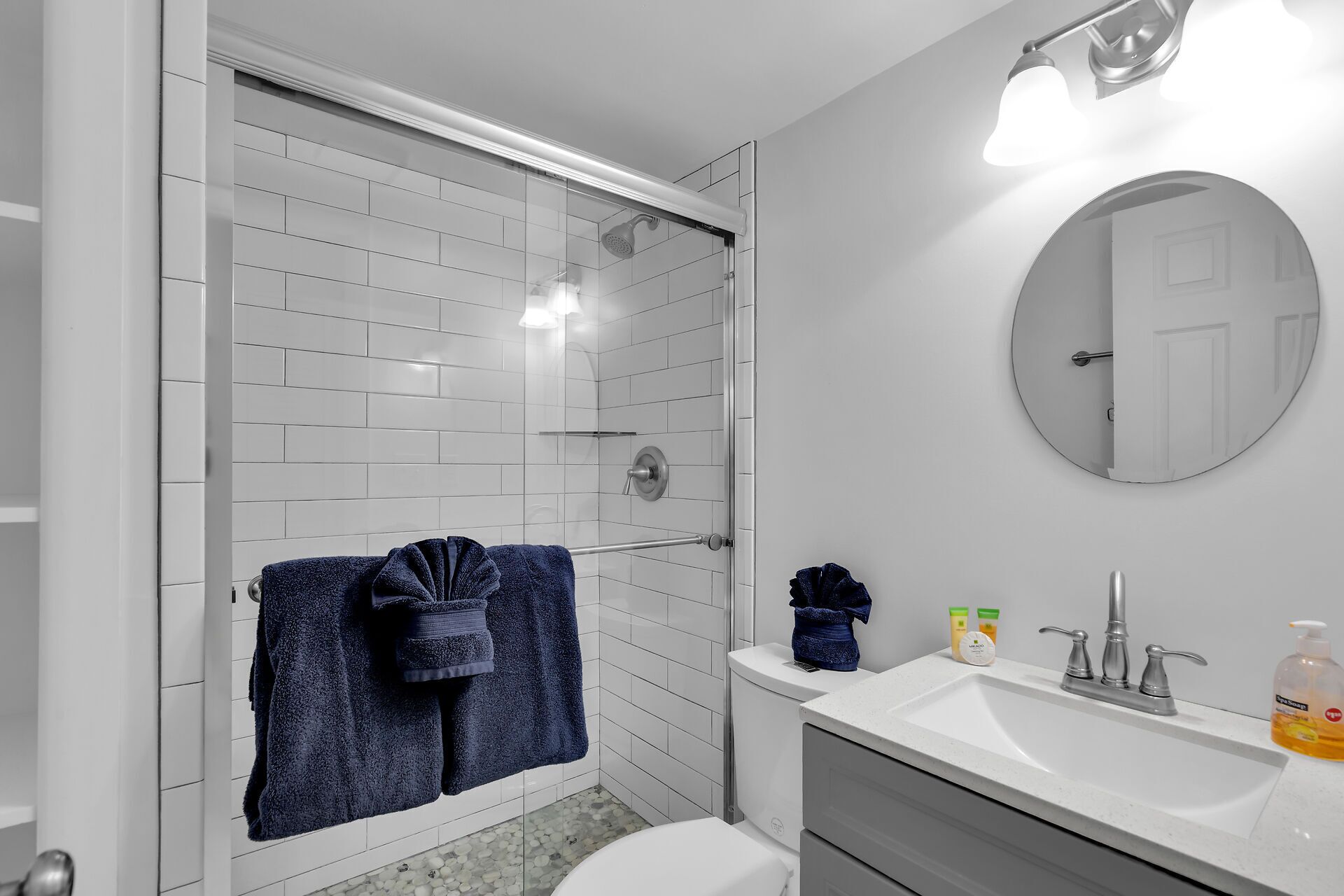 A modern bathroom with a glass shower enclosure, white tiled walls, a round mirror, and a white sink with toiletries.