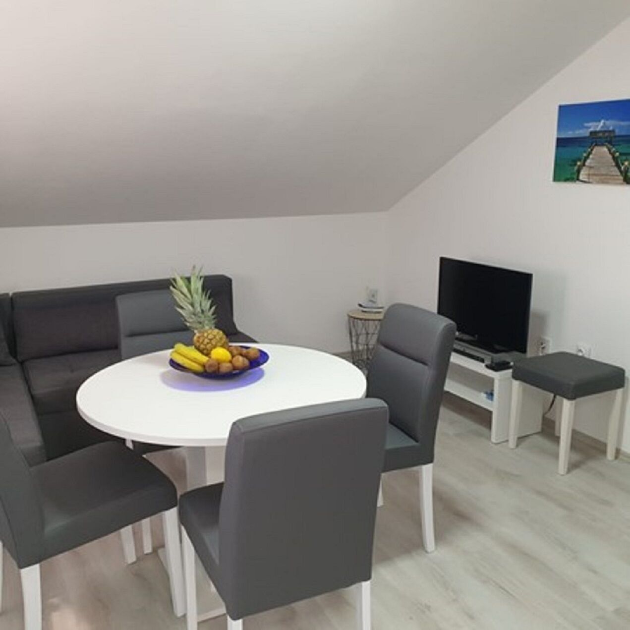 Ein modernes Wohnzimmer mit einem runden weißen Tisch, grauen Sofas, einem Flachbildfernseher und einem kleinen Beistelltisch.