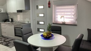A compact kitchen with white cabinets, a stove, and a sink. There's a dining table with chairs and a fruit bowl. A window with a plant is visible. - App. 80 meters from the sea (Vir)