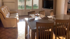 A living room with a sofa, a dining table with chairs, a TV, and a sliding glass door leading to a garden.