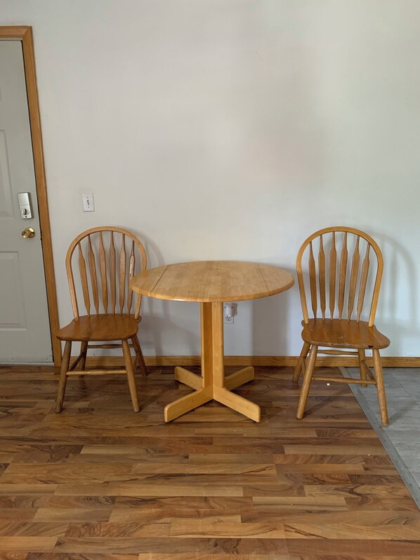 Una mesa redonda de madera con una base central de pedestal, flanqueada por dos sillas de madera con respaldo de listones. La habitación tiene un piso de parquet de madera y una pared blanca lisa con una puerta a la izquierda.