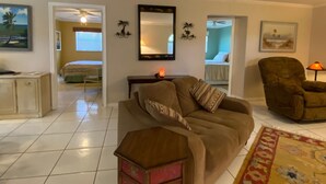 A living room with a sofa, a chair, a small table, and a mirror on the wall. There is a bed in the background and a window with blinds. - Vintage Vero Bungalow (Vero Beach)