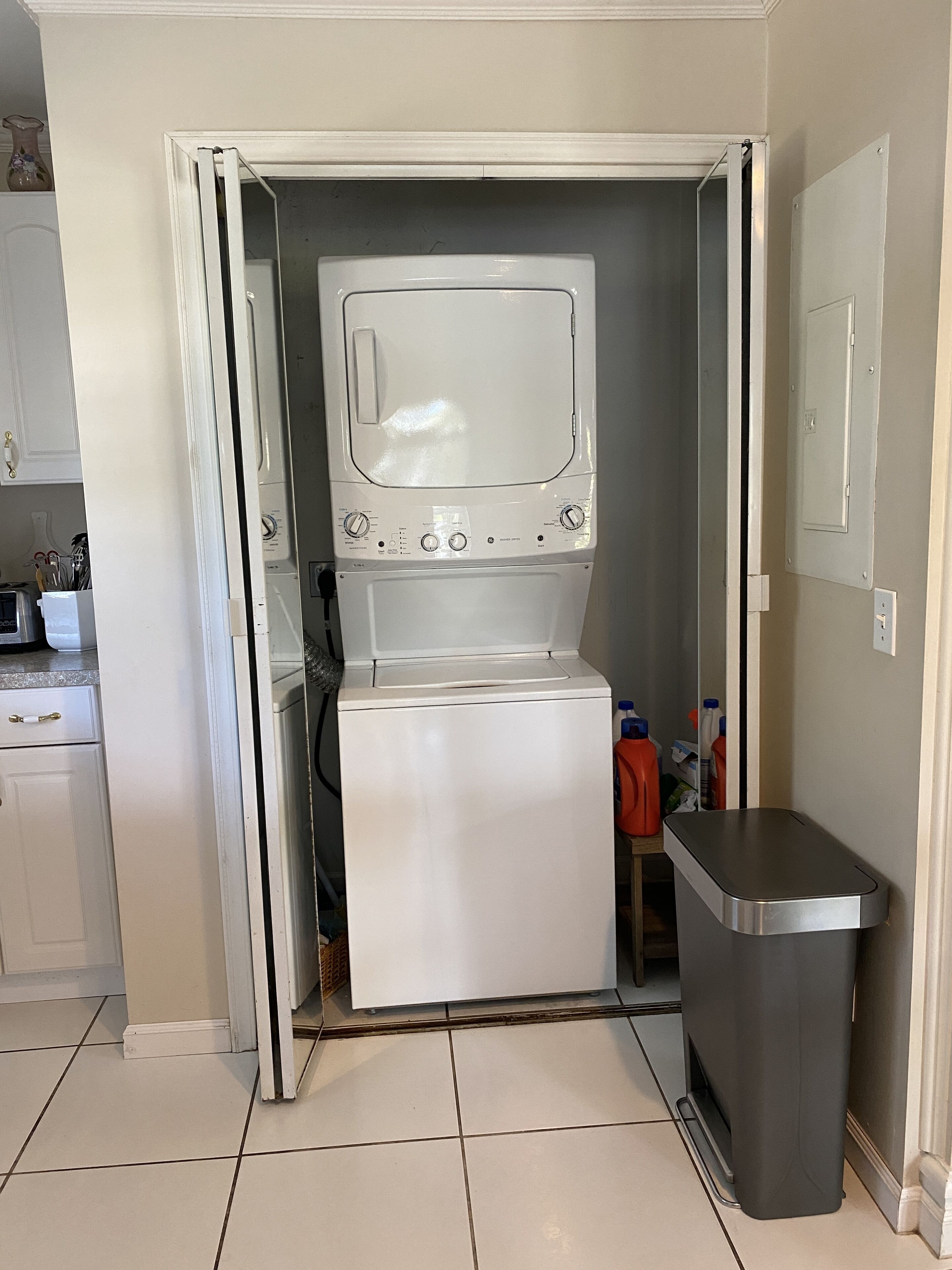 Une petite buanderie équipée d’un lave-linge à chargement frontal, d’une poubelle et d’une petite étagère contenant des produits de nettoyage.