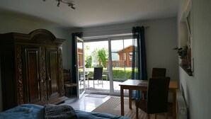 Een kamer met een bed, een houten kledingkast, een eettafel met stoelen en een schuifdeur die leidt naar een buitenruimte met uitzicht op een gebouw en groen.