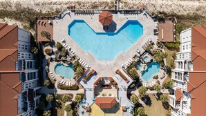 Eine Vogelperspektive zeigt ein Resort mit mehreren Schwimmbecken, darunter ein zentral gelegenes, herzförmiges Becken, umgeben von Liegebereichen und Palmen.