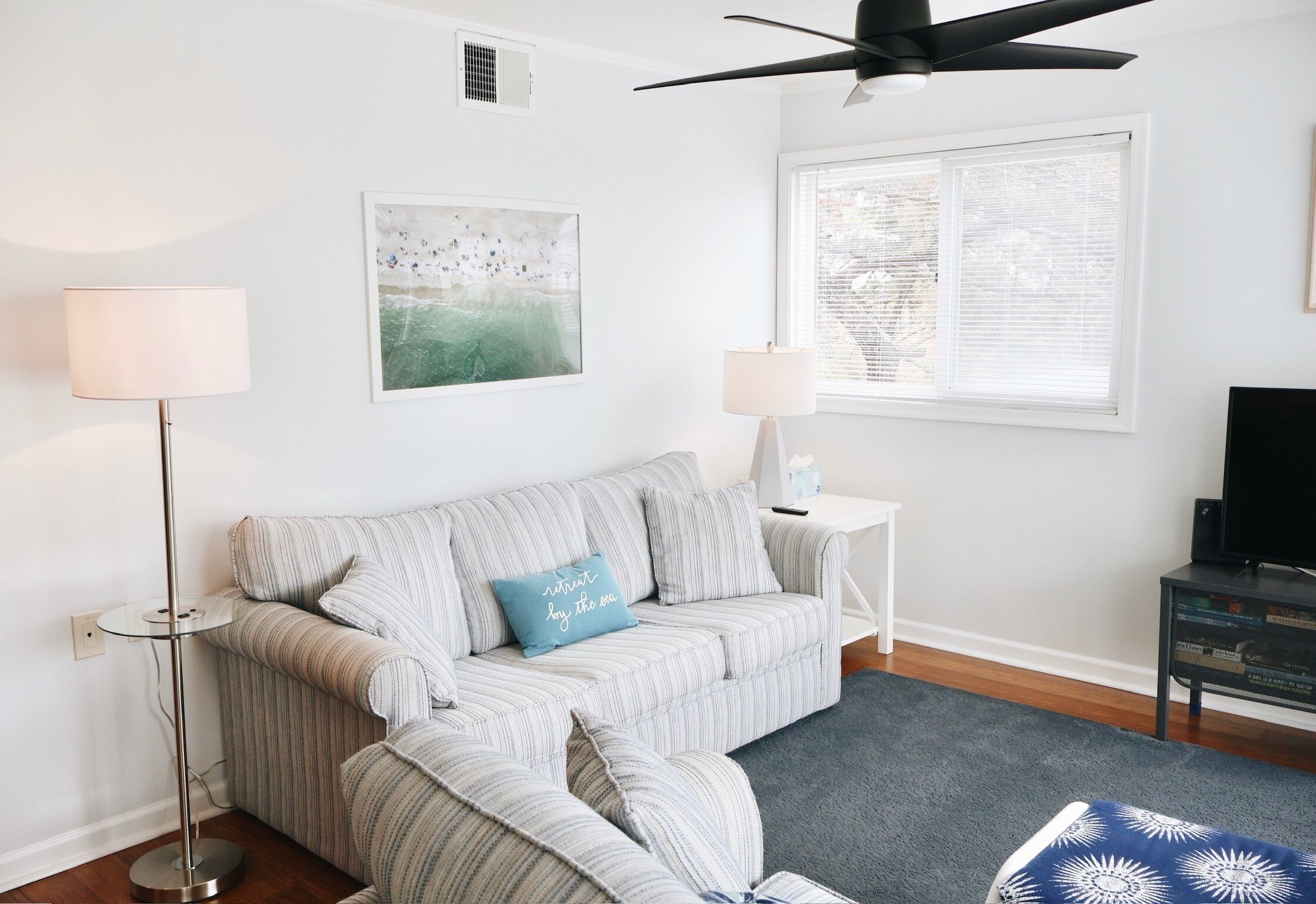 A living room with a striped sofa, two side tables, a ceiling fan, a large window with blinds, and a television on a stand.