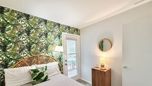 Un dormitorio con cama, una silla de mimbre, una mesita de noche con lámpara, un espejo y una puerta que da a un balcón.