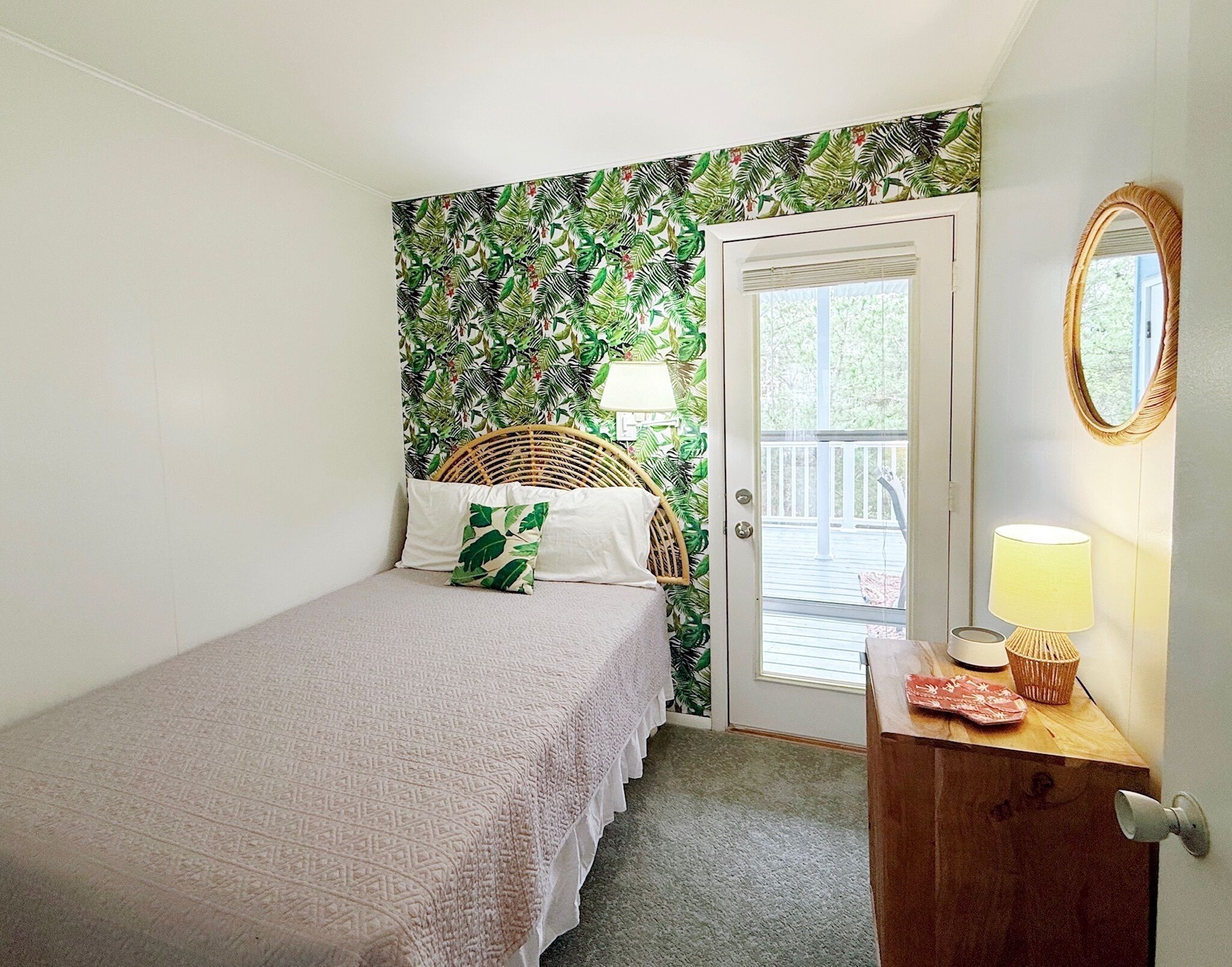 A bedroom with a bed, a wicker chair, a door leading to a balcony, a wooden dresser, and a mirror.
