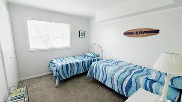 Une chambre avec deux lits simples, une petite table, une lampe, une fenêtre avec des stores et une étagère fixée au mur, sur laquelle est posée une plaque nominative.