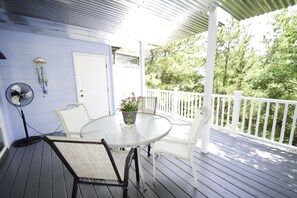 Une terrasse extérieure couverte, dotée d’une table ronde en verre et de chaises blanches, d’une porte blanche et d’une clôture blanche, qui offre une vue sur une zone boisée.