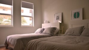 Ein Schlafzimmer mit zwei Betten, einem weißen Kopfteil und zwei Fenstern mit Blick auf Palmen.
