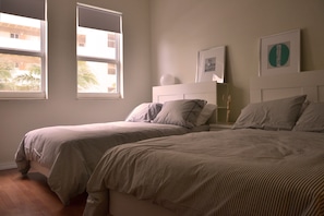 A bedroom with two beds, a white headboard, and two windows with views of palm trees.