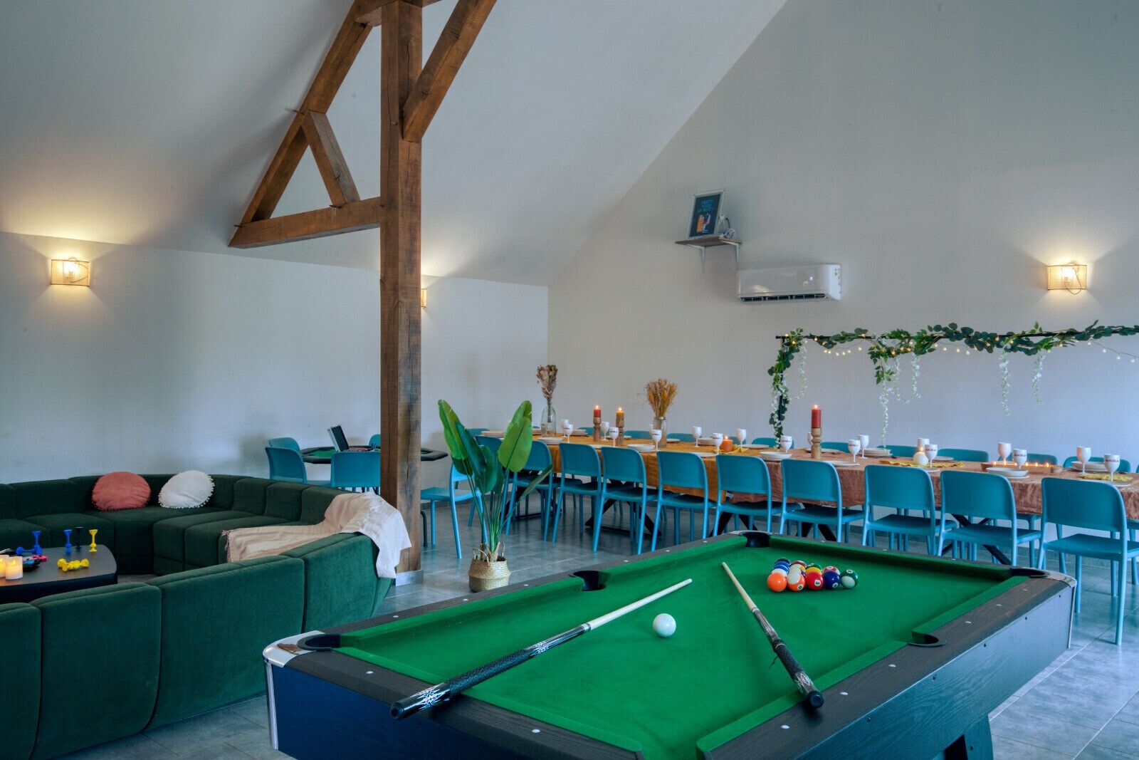 A pool table in a room with a high ceiling and exposed wooden beams. There are blue chairs around a table set for dining, and a green sofa with cushions.