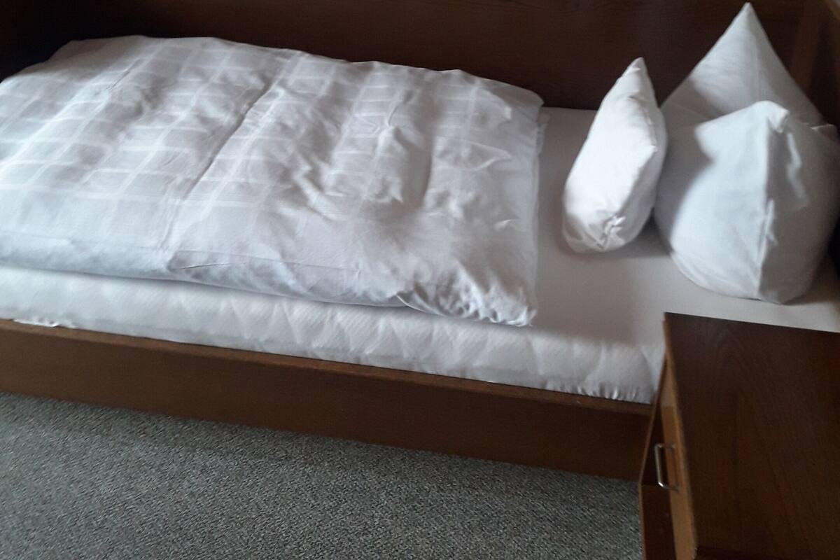 Un lit simple avec une tête de lit en bois, une literie blanche et un sol recouvert d’un tapis gris.