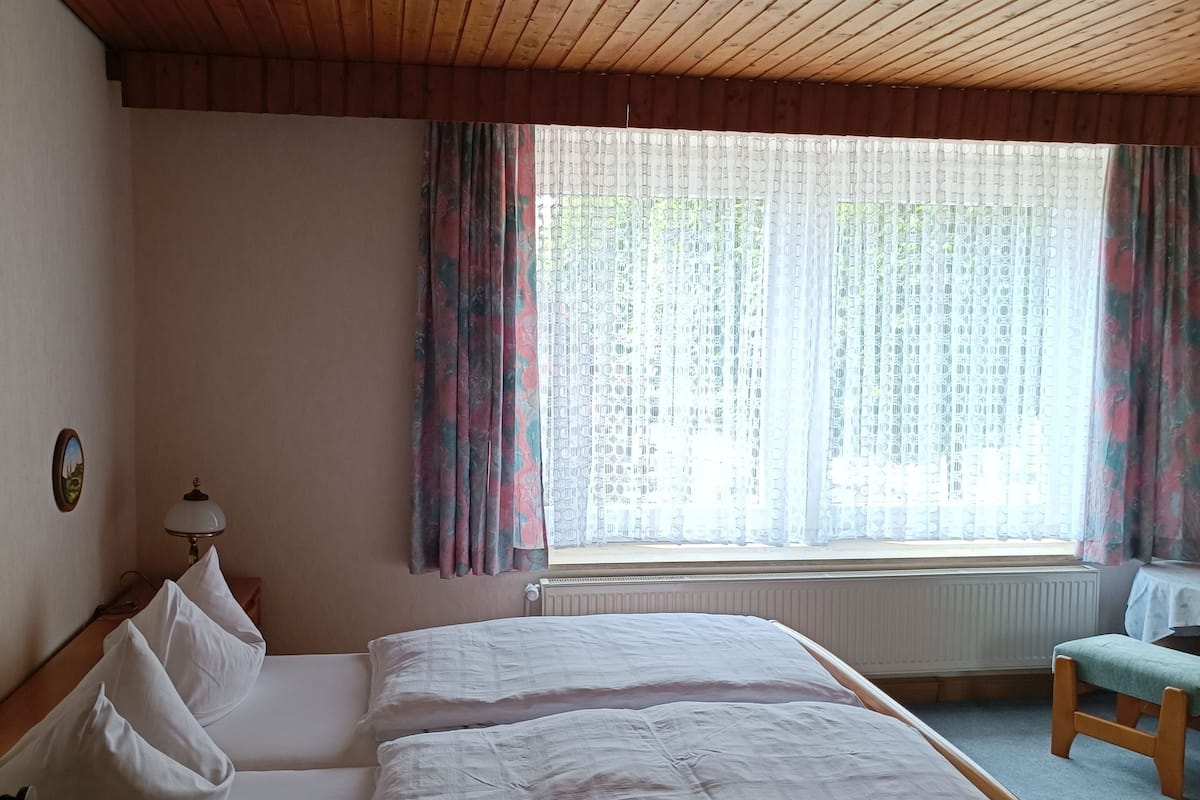Une pièce avec un plafond en bois, un lit avec une literie blanche, une table de chevet, une lampe, une fenêtre avec des rideaux et un radiateur.