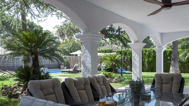 An outdoor patio with a glass table, cushioned chairs, and a pool in the background.