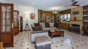 A spacious living room with a large sectional sofa, a coffee table, and a fireplace. There is a wooden bookshelf and a floor lamp.