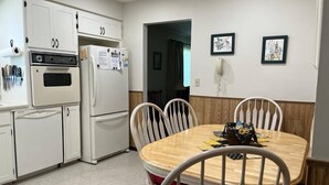 Una cocina con armarios blancos, una estufa y un refrigerador. Hay una mesa de comedor con sillas y un tapete rojo.