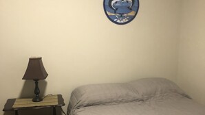 Un dormitorio con una cama, una mesita de noche con una lámpara, un reloj de pared y una pequeña estantería con libros.