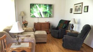 Una sala de estar con sofá, dos sillones, una mesa de centro de madera, televisor montado en la pared y un mueble de madera.