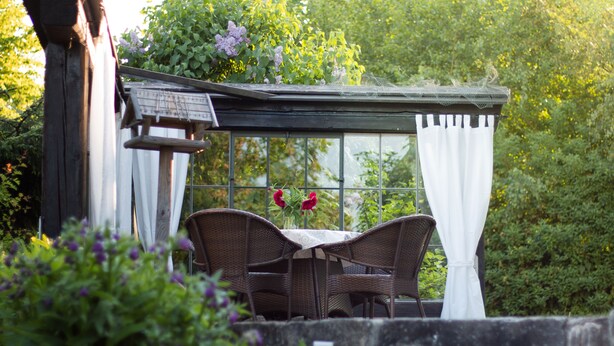 A conservatory with wicker furniture and a table, surrounded by greenery and flowers.
