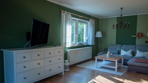 A living room with a television on a white dresser, a grey sofa, a wooden coffee table, and a window with curtains.