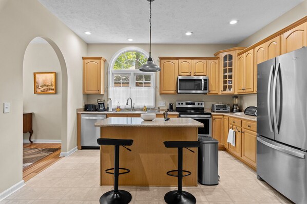 A modern kitchen with wooden cabinets, stainless steel appliances, a large island with bar stools, and a window with a view of greenery.
