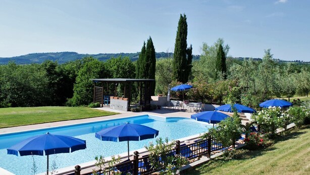 Una piscina con sombrillas azules, una pérgola y vistas del entorno verde.