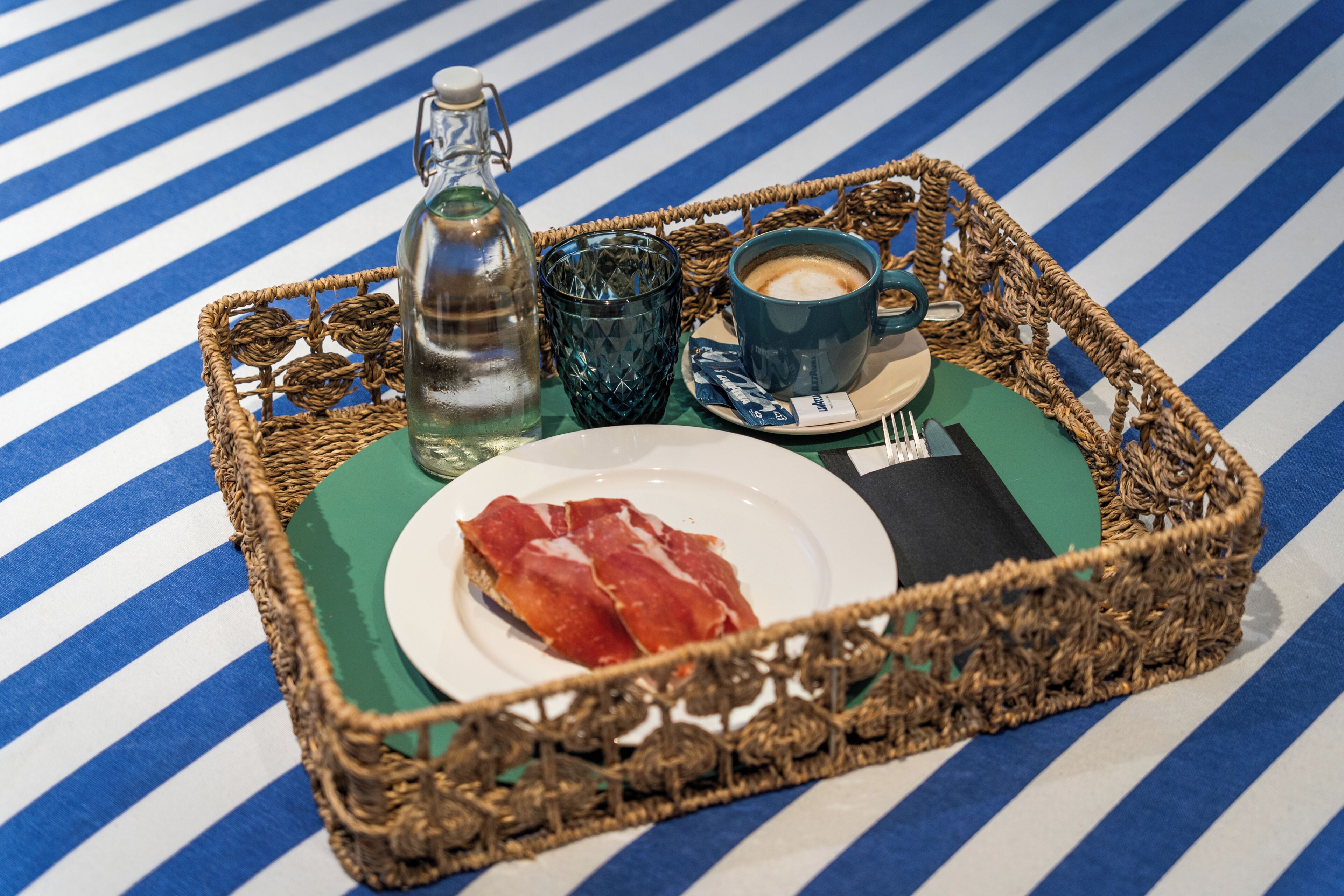 A tray with a bottle, a glass, a cup, a plate with food, and a napkin on a striped surface.